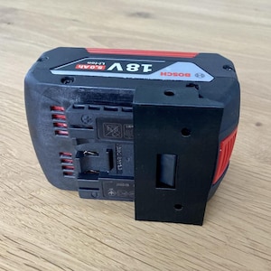 Op de afbeelding: Een zwarte en rode Bosch 18V Li-ion 6.0Ah accu met een zwarte montagebeugel. De accu heeft een rechthoekige vorm met rode accenten en het Bosch-logo. De beugel heeft drie gaten voor montage.