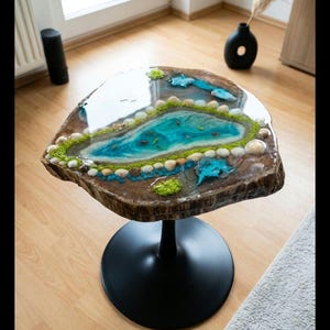 Könnte beinhalten: Einzigartiger, handgefertigter Couchtisch mit einem Harzaufsatz, der eine Seelandschaft darstellt. Der Tisch hat eine natürliche Holzkante, ein blau-grünes See-Design mit kleinen Fischen und wird von einem schwarzen Metallsockel getragen. Der Tisch hat einen Durchmesser von ca. 76 cm.