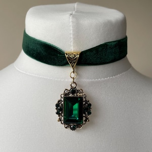 May include: A dark green velvet choker necklace featuring a gold-toned metal connector and a central pendant. The pendant consists of a large, rectangular emerald-coloured faceted stone surrounded by an ornate filigree frame with small dark accents.
