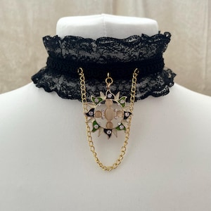 Puede incluir: Collar gargantilla de encaje negro con colgante dorado. El colgante es un diseño de sol con una piedra ovalada central, rodeada de acentos verdes, blancos y transparentes. El collar tiene una delicada cadena dorada.