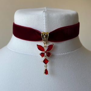 May include: A deep red velvet choker necklace featuring a gold-toned metal bail and a dangling pendant. The pendant consists of a red butterfly-shaped crystal arrangement, a square red crystal, and a teardrop-shaped red crystal at the base.