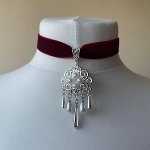 May include: A burgundy velvet choker necklace with a silver pendant. The pendant has a floral design with faux pearls and dangling teardrop charms. The necklace is displayed on a white mannequin.