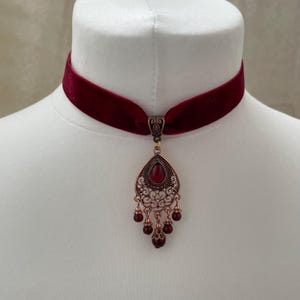 Könnte beinhalten: Burgunderfarbenes Samt-Choker-Halsband mit einem verzierten kupferfarbenen Anhänger. Der Anhänger hat einen tropfenförmigen roten Edelstein und hängende rote Perlen, die einen Vintage-Look erzeugen. Die Halskette wird auf einer weißen Schaufensterpuppe präsentiert.