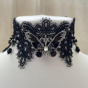 Op de afbeelding: Zwarte kanten choker ketting met een vlinderhanger. De vlinder is zwart met zilverkleurige details en een centrale heldere edelsteen. De choker heeft kralen accenten en een delicate, geschulpte kanten rand.