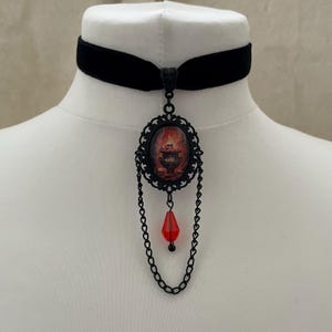 Könnte beinhalten: Ein schwarzes Samt-Halsband mit einem ovalen Anhänger, der eine Grafik mit dunkelroten Flammen und einem Totenkopf zeigt. Der Anhänger ist in schwarzes Filigranmetall gefasst und mit zwei dünnen schwarzen Ketten sowie einer roten Tropfenperle versehen.