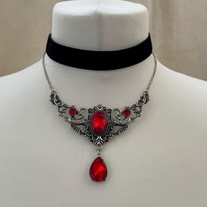 May include: A black velvet choker necklace featuring a silver-toned metal centerpiece with intricate scrollwork, three round red stones, and a single dangling teardrop-shaped red stone pendant suspended from a delicate silver-toned chain.