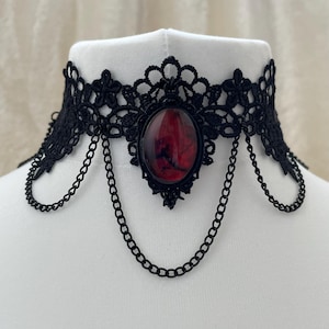 Puede incluir: Collar gargantilla de encaje negro con un colgante ovalado central con un diseño rojo y negro. La gargantilla tiene un delicado patrón de encaje floral y está adornada con detalles de cadena negra colgantes. El collar se muestra en un maniquí blanco.