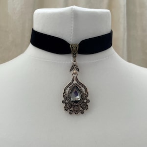 以下が含まれることがあります： 黒いベルベットのチョーカーネックレス。アンティーク調の装飾的なペンダントが付いています。ペンダントは、小さなクリスタルと装飾的な金属細工に囲まれた、大きな洋梨型の灰色のクリスタルが特徴です。ネックレスは白いマネキンに飾られています。