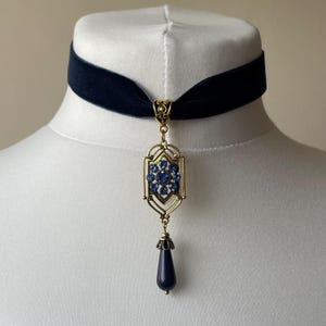 Könnte beinhalten: Ein marineblauer Samt-Choker-Halskette mit einem goldfarbenen Anhänger. Der Anhänger hat ein blaues Edelstein-Design und einen tropfenförmigen Anhänger. Die Halskette wird auf einer weißen Schaufensterpuppe präsentiert.