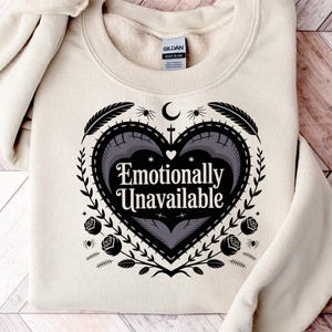 May include: A sand-colored sweatshirt with the text "Emotionally Unavailable" inside a heart-shaped design. The design includes feathers, roses, and a crescent moon. The sweatshirt is folded on a wooden surface.