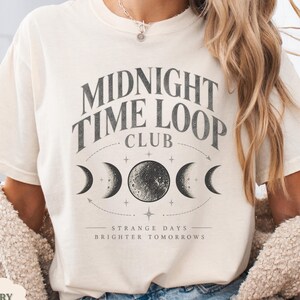 May include: An ivory short-sleeve t-shirt featuring a dark gray graphic with the text MIDNIGHT TIME LOOP CLUB and STRANGE DAYS BRIGHTER TOMORROWS. The design includes a central full moon flanked by crescent moon phases and small stars.