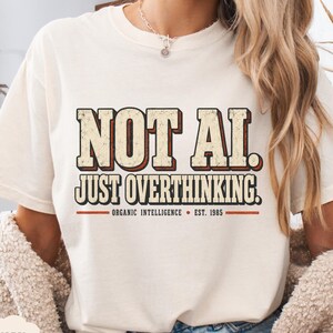 May include: An ivory short-sleeve t-shirt featuring the text "NOT AI. JUST OVERTHINKING." in a distressed, block-style font with red and black outlines. Below the main text, it reads "ORGANIC INTELLIGENCE • EST. 1985" with horizontal red lines.