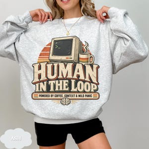 May include: An ash gray crewneck sweatshirt featuring a retro-style graphic of a vintage computer monitor and a coffee mug. The text reads "HUMAN IN THE LOOP" and "POWERED BY COFFEE, CONTEXT & MILD PANIC" above a small brain icon with radiating lines.