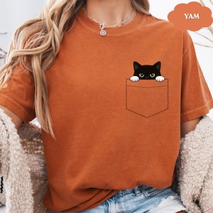 A woman wearing an orange t-shirt with a black cat graphic peeking out of a pocket.