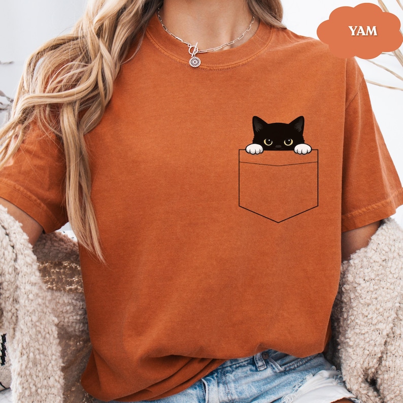 A woman wearing an orange t-shirt with a black cat graphic peeking out of a pocket.