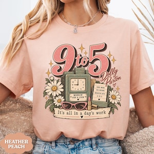 May include: A heather peach t-shirt featuring a graphic print with the text 9 to 5 Dolly Parton and It's all in a day's work. The design includes a vintage clock, a notepad with text, pink cat-eye glasses, a lipstick tube, and white daisies.