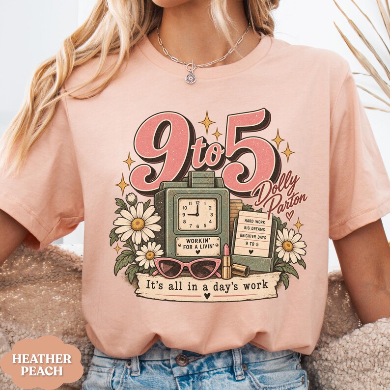 May include: A heather peach t-shirt featuring a graphic print with the text 9 to 5 Dolly Parton and It's all in a day's work. The design includes a vintage clock, a notepad with text, pink cat-eye glasses, a lipstick tube, and white daisies.
