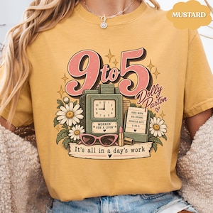 May include: A mustard yellow t-shirt featuring a graphic print with the text 9 to 5, Dolly Parton, and It's all in a day's work. The design includes a vintage clock, daisies, pink sunglasses, a lipstick tube, and a notepad with text.