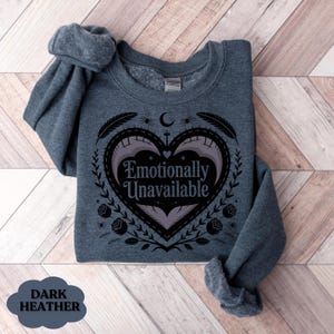 Puede incluir: Sudadera gris jaspeado oscuro con el texto "Emotionally Unavailable" dentro de un dise&ntilde;o en forma de coraz&oacute;n. El dise&ntilde;o incluye elementos florales y celestiales. Las mangas de la sudadera est&aacute;n enrolladas.