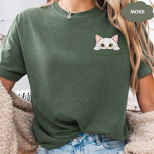A woman wearing a green t-shirt with a white cat graphic peeking out of a pocket.