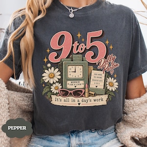 May include: A charcoal gray t-shirt featuring a graphic with the text 9 to 5 Dolly Parton and It's all in a day's work. The design includes a vintage clock, daisies, pink sunglasses, a lipstick tube, and a sign listing work-related phrases.