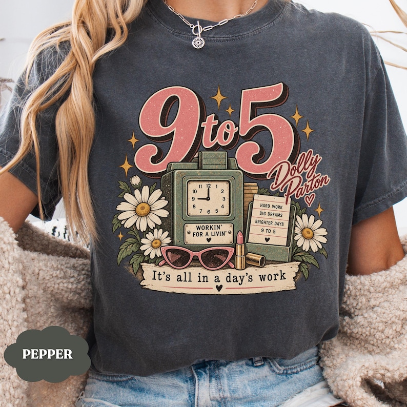 May include: A charcoal gray t-shirt featuring a graphic with the text 9 to 5 Dolly Parton and It's all in a day's work. The design includes a vintage clock, daisies, pink sunglasses, a lipstick tube, and a sign listing work-related phrases.