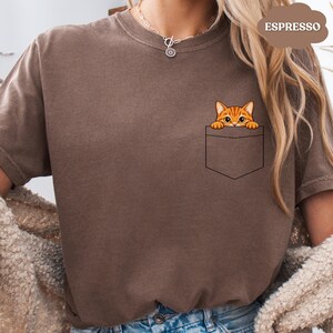 A person wearing a brown t-shirt with a small orange cat peeking out of a pocket.