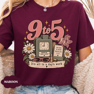 May include: A maroon t-shirt featuring a graphic print with the text 9 to 5, Dolly Parton, and It's all in a day's work. The design includes a vintage clock, a sign with text, pink cat-eye glasses, a lipstick tube, and white daisy flowers.