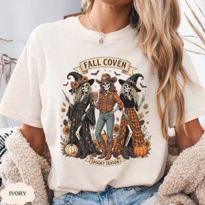 May include: An ivory t-shirt featuring a graphic of three skeletons dressed in fall attire with the text FALL COVEN and SPOOKY SEASON. The skeletons wear hats, plaid shirts, and skirts, surrounded by sunflowers, pumpkins, and bats in a fall theme.