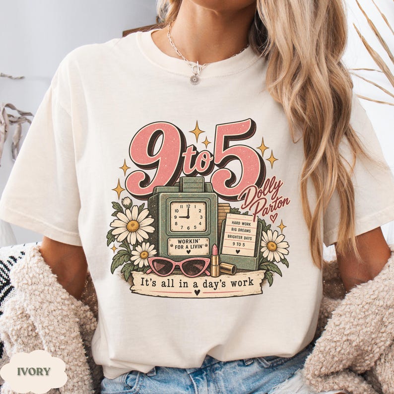 May include: An ivory t-shirt featuring a graphic print with the text 9 to 5 Dolly Parton and It's all in a day's work. The design includes a green clock, pink sunglasses, a lipstick tube, and white daisies with yellow centers.
