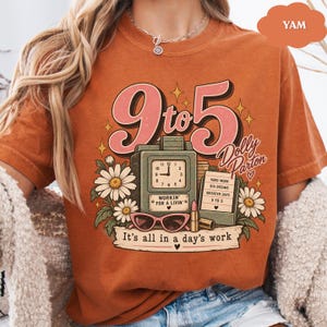 May include: A burnt orange t-shirt featuring a graphic print with the text 9 to 5, Dolly Parton, and It's all in a day's work. The design includes a vintage time clock, daisies, pink sunglasses, a lipstick tube, and a notepad with text.