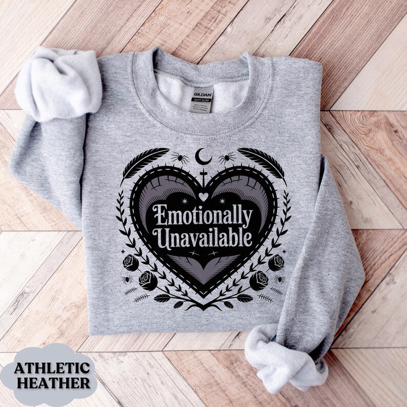 Puede incluir: Sudadera gris jaspeado con el texto "Emotionally Unavailable" dentro de un dise&ntilde;o en forma de coraz&oacute;n. El dise&ntilde;o incluye plumas, ara&ntilde;as, rosas y una luna creciente. La sudadera est&aacute; doblada sobre una superficie de madera.