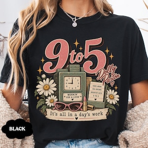 May include: A black t-shirt featuring a graphic print with the text 9 to 5, Dolly Parton, and It's all in a day's work. The design includes a green time clock, white daisies, pink cat-eye sunglasses, a lipstick tube, and a notepad with text.