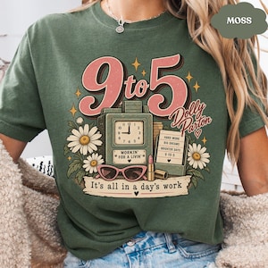 May include: A moss green t-shirt featuring a graphic with the text 9 to 5, Dolly Parton, Workin' for a livin', and It's all in a day's work. The design includes a clock, a sign, daisies, a lipstick tube, and pink cat-eye glasses.