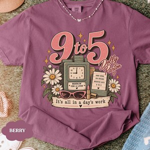 May include: A berry-colored t-shirt featuring a graphic print with the text 9 to 5, Dolly Parton, and It's all in a day's work. The design includes a vintage clock, a notepad, sunglasses, lipstick, and white daisies with green leaves.