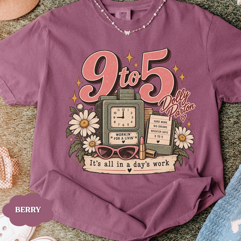 May include: A berry-colored t-shirt featuring a graphic print with the text 9 to 5, Dolly Parton, and It's all in a day's work. The design includes a vintage clock, a notepad, sunglasses, lipstick, and white daisies with green leaves.