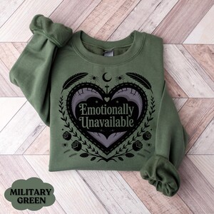 Puede incluir: Una sudadera verde militar con el texto "Emotionally Unavailable" dentro de un dise&ntilde;o en forma de coraz&oacute;n. El dise&ntilde;o incluye espinas, rosas y una luna creciente. La sudadera est&aacute; doblada sobre una superficie de madera.