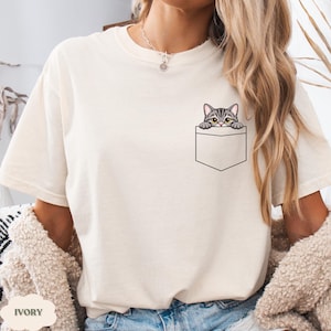 A woman wearing a white t-shirt with a cat graphic peeking out of a pocket.