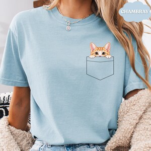 A woman wearing a light blue t-shirt with a cat graphic peeking out of a pocket.
