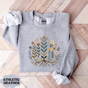 May include: Athletic heather gray sweatshirt featuring a floral design with various flowers and leaves in orange, yellow, and green. The design is centered on the chest. The text "ATHLETIC HEATHER" is at the bottom.