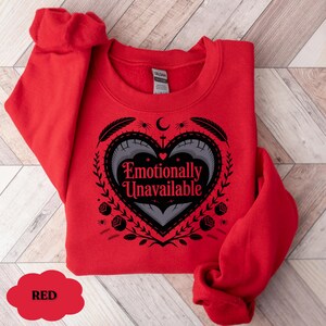 Puede incluir: Una sudadera roja con el texto "Emotionally Unavailable" dentro de un dise&ntilde;o en forma de coraz&oacute;n. El dise&ntilde;o incluye plumas, rosas y una luna creciente. La sudadera es de material suave.