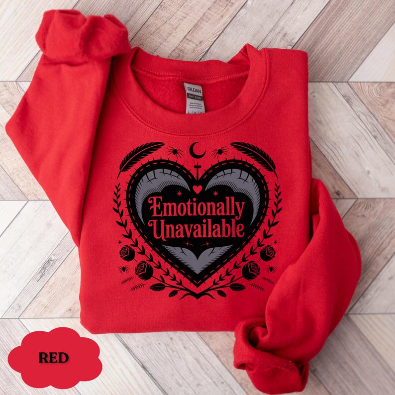 Puede incluir: Una sudadera roja con el texto "Emotionally Unavailable" dentro de un dise&ntilde;o en forma de coraz&oacute;n. El dise&ntilde;o incluye plumas, rosas y una luna creciente. La sudadera es de material suave.