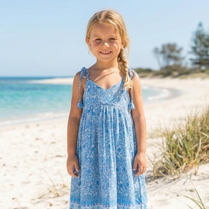 Op de afbeelding: Een jong meisje staat op een zandstrand en draagt een blauwe jurk met bloemenprint. De jurk heeft een V-hals, strikbandjes en een zwierige rok. De jurk heeft een wit en blauw bloemenpatroon. Het meisje heeft blond haar en lacht.