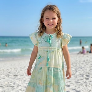 以下が含まれることがあります： 砂浜に立つ、パステルカラーのドレスを着た少女。ドレスは、ティアードスカートとフレアスリーブで、緑、黄、青の色合いの水彩画のような模様が特徴です。ネックラインにはタッセルのディテールがあります。背景には海と青い空が広がっています。