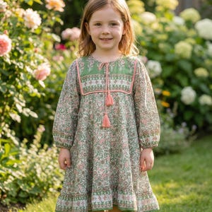 Puede incluir: Una niña con un vestido de manga larga con estampado floral en tonos verdes y rosas. El vestido tiene un canesú verde con ribete y borlas rosas. La niña sonríe en un jardín.