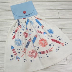 May include: A white kitchen towel with a blue top and red button. The towel is decorated with red, white, and blue fireworks and stars, perfect for a patriotic theme. The towel is designed to hang.