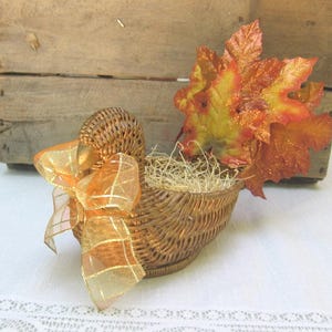 May include: A woven wicker turkey-shaped basket featuring a gold-toned beak, a sheer orange plaid ribbon tied around the neck, and a tail made of artificial autumn leaves in shades of red, orange, and yellow. The center is filled with straw.