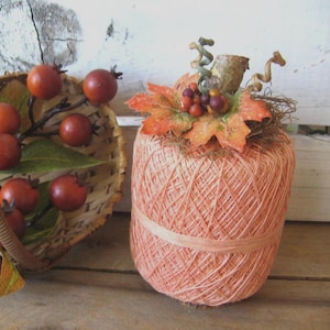 May include: A decorative pumpkin crafted from peach-colored string wrapped in a cylindrical shape, topped with artificial orange maple leaves, small dark berries, and a textured stem, positioned beside a woven basket filled with faux red berries.