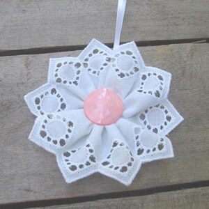 May include: A white fabric ornament shaped like a flower with scalloped edges and eyelet lace detailing. The center features a smooth, round, light pink button. A thin white ribbon is attached to the top for hanging the decorative piece.