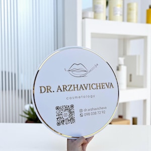 Puede incluir: Un letrero redondo blanco con borde dorado. Muestra el texto "DR. ARZHAVICHEVA cosmetology", un gráfico de labios y jeringa, un código QR e información de contacto.